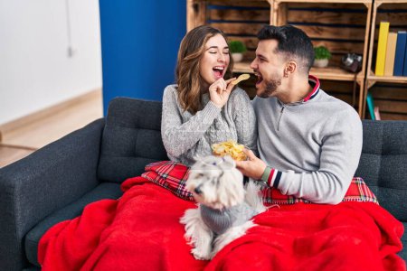 Foto de Man and woman eating chips potatoes sitting on sofa with dog at home - Imagen libre de derechos