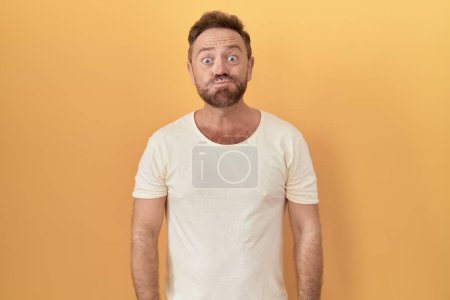 Téléchargez les photos : Middle age man with beard standing over yellow background puffing cheeks with funny face. mouth inflated with air, crazy expression. - en image libre de droit