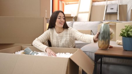 Foto de Joven hermosa mujer hispana sonriendo confiada desempacando caja de cartón en nuevo hogar - Imagen libre de derechos