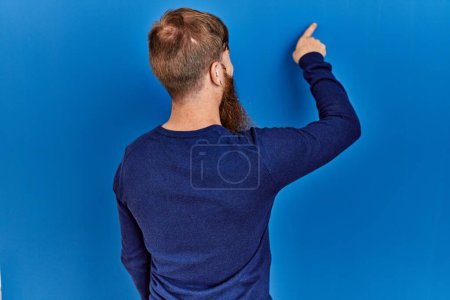 Photo for Redhead man with long beard wearing casual blue sweater over blue background posing backwards pointing ahead with finger hand - Royalty Free Image