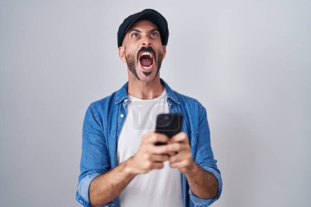 Foto de Hombre hispano con barba usando teléfono inteligente escribiendo mensaje enojado y loco gritando frustrado y furioso, gritando con ira mirando hacia arriba. - Imagen libre de derechos