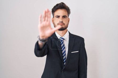 Foto de Young hispanic man with tattoos wearing business suit and tie doing stop sing with palm of the hand. warning expression with negative and serious gesture on the face. - Imagen libre de derechos