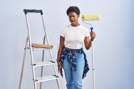 Foto de Mujer afroamericana sosteniendo rodillo pintor enojado y loco gritando frustrado y furioso, gritando con ira. rabia y concepto agresivo. - Imagen libre de derechos