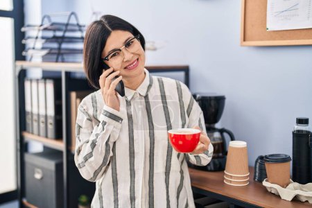 Photo for Young chinese woman business worker talking on the martphone and drinking coffee at office - Royalty Free Image