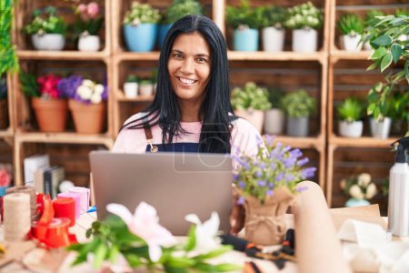 Foto de Florista hispana de mediana edad sonriendo confiada usando laptop en floristería - Imagen libre de derechos