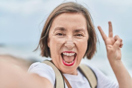 Téléchargez les photos : Femme d'âge moyen souriant confiant faire selfie par caméra au bord de la mer - en image libre de droit