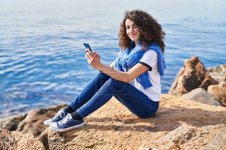 Foto de Mujer hispana joven sonriendo confiada usando smartphone en la playa - Imagen libre de derechos