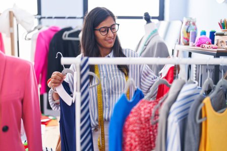 Photo for Young beautiful hispanic woman tailor smiling confident holding clothes on rack at clothing factory - Royalty Free Image