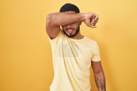 Foto de Joven hombre hispano de pie sobre fondo amarillo cubriendo los ojos con el brazo sonriente alegre y divertido. concepto ciego. - Imagen libre de derechos