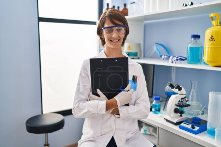 Foto de Joven hermosa mujer hispana científica sonriendo confiada sosteniendo portapapeles en el laboratorio - Imagen libre de derechos