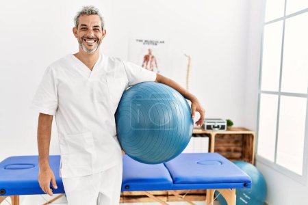 Foto de Middle age grey-haired man physiotherapist smiling confident holding yoga mat at rehab clinic - Imagen libre de derechos