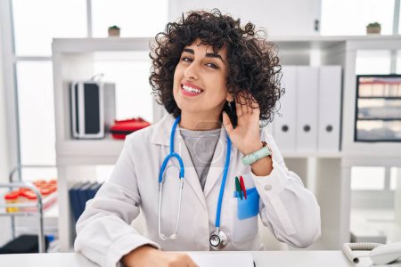 Photo for Young brunette woman with curly hair wearing doctor uniform and stethoscope smiling with hand over ear listening and hearing to rumor or gossip. deafness concept. - Royalty Free Image
