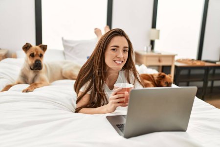 Téléchargez les photos : Jeune femme hispanique utilisant un ordinateur portable et buvant du café couché sur le lit avec des chiens dans la chambre - en image libre de droit