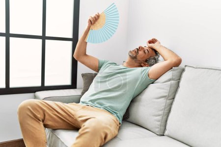 Téléchargez les photos : Moyen âge homme aux cheveux gris en utilisant un ventilateur assis sur le canapé à la maison - en image libre de droit