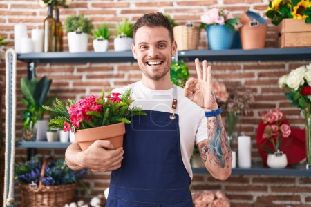 Foto de Joven hombre hispano trabajando en floristería sosteniendo maceta haciendo ok signo con los dedos, sonriendo gesto amistoso excelente símbolo - Imagen libre de derechos