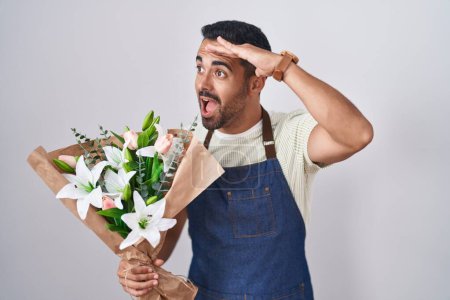 Foto de Hombre hispano con barba trabajando como florista muy feliz y sonriente mirando lejos con la mano sobre la cabeza. concepto de búsqueda. - Imagen libre de derechos