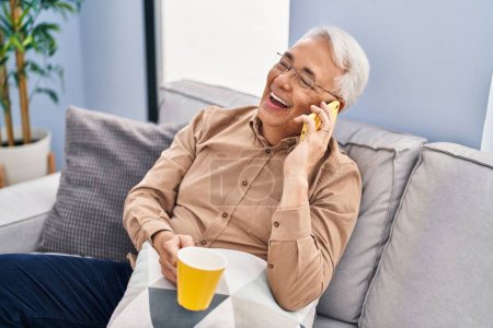 Téléchargez les photos : Homme âgé boire du café et parler sur le smartphone à la maison - en image libre de droit
