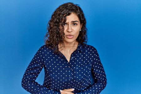 Photo for Young brunette woman with curly hair wearing casual clothes over blue background with hand on stomach because indigestion, painful illness feeling unwell. ache concept. - Royalty Free Image