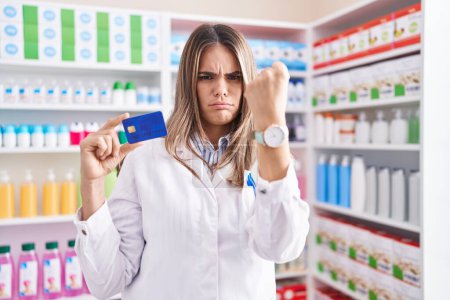Foto de Mujer joven hispana que trabaja en farmacia con tarjeta de crédito molesta y frustrada gritando de rabia, gritando loca de rabia y levantando la mano - Imagen libre de derechos