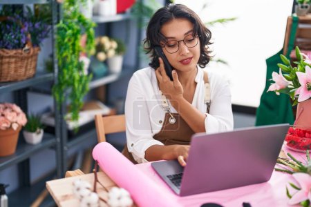 Photo for Young beautiful hispanic woman florist talking on smartphone using laptop at flower shop - Royalty Free Image