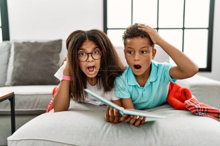 Photo for Two siblings lying on the sofa reading a book crazy and scared with hands on head, afraid and surprised of shock with open mouth - Royalty Free Image