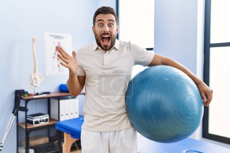 Foto de Handsome hispanic man holding pilates ball at rehabilitation clinic celebrating victory with happy smile and winner expression with raised hands - Imagen libre de derechos