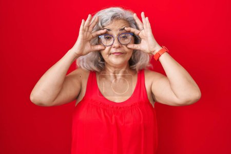 Foto de Middle age woman with grey hair standing over red background trying to open eyes with fingers, sleepy and tired for morning fatigue - Imagen libre de derechos