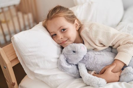 Photo for Adorable blonde girl hugging rabbit doll lying on bed at bedroom - Royalty Free Image