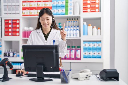 Téléchargez les photos : Jeune femme chinoise travaillant à la pharmacie pharmacie portant un casque souriant avec une idée ou une question pointant du doigt avec un visage heureux, numéro un - en image libre de droit
