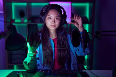 Téléchargez les photos : Young asian woman playing video games with smartphone smiling and confident gesturing with hand doing small size sign with fingers looking and the camera. measure concept. - en image libre de droit