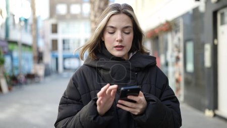 Téléchargez les photos : Jeune femme blonde utilisant un smartphone avec une expression sérieuse à la rue - en image libre de droit