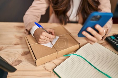 Téléchargez les photos : Jeune femme hispanique e-commerce travailleur d'affaires en utilisant smartphone et l'écriture sur le paquet au bureau - en image libre de droit