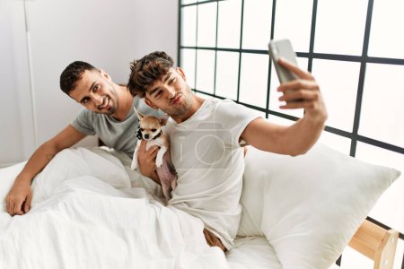 Photo for Two hispanic men couple make selfie by the smartphone sitting on bed with chihuahua at bedroom - Royalty Free Image
