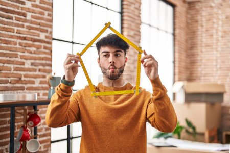 Foto de Hispanic man with beard moving to a new home holding ruler making fish face with mouth and squinting eyes, crazy and comical. - Imagen libre de derechos