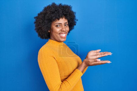 Foto de Mujer negra con el pelo rizado de pie sobre fondo azul señalando a un lado con las manos abiertas palmeras mostrando espacio de copia, presentando anuncio sonriente emocionado feliz - Imagen libre de derechos