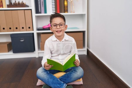 Foto de Adorable niño hispano estudiante sonriendo confiado lectura libro en biblioteca escuela - Imagen libre de derechos