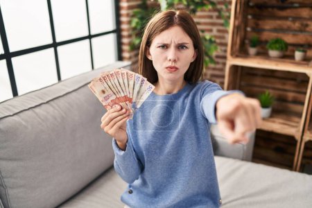 Téléchargez les photos : Young brunette woman holding colombian pesos pointing with finger to the camera and to you, confident gesture looking serious - en image libre de droit