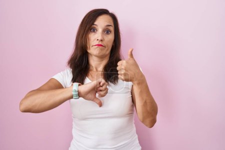 Foto de Middle age brunette woman standing over pink background doing thumbs up and down, disagreement and agreement expression. crazy conflict - Imagen libre de derechos