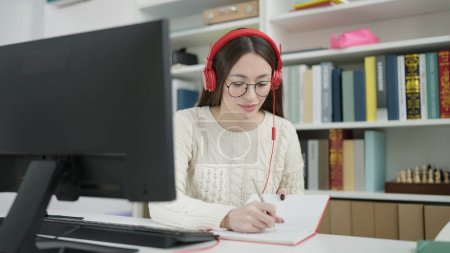 Foto de Joven hermosa estudiante hispana usando computadora y auriculares escribiendo en un cuaderno en la universidad de la biblioteca - Imagen libre de derechos