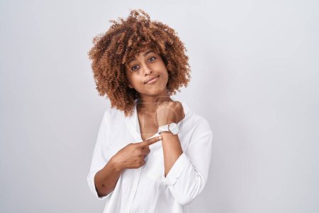 Foto de Young hispanic woman with curly hair standing over white background in hurry pointing to watch time, impatience, looking at the camera with relaxed expression - Imagen libre de derechos