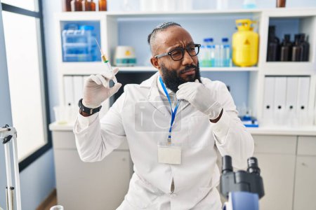 Foto de African american man working at scientist laboratory holding syringe serious face thinking about question with hand on chin, thoughtful about confusing idea - Imagen libre de derechos
