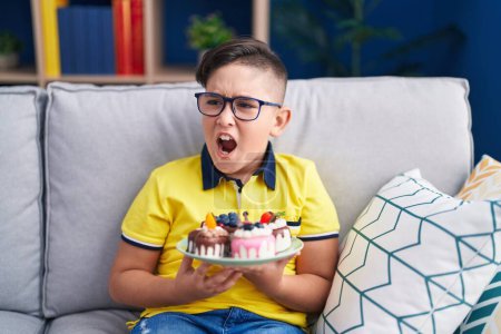Foto de Joven niño hispano sosteniendo dulces de pastel enojado y loco gritando frustrado y furioso, gritando con ira. rabia y concepto agresivo. - Imagen libre de derechos