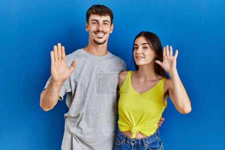 Téléchargez les photos : Jeune couple hispanique debout ensemble sur fond bleu renonçant à dire bonjour heureux et souriant, geste de bienvenue amical - en image libre de droit
