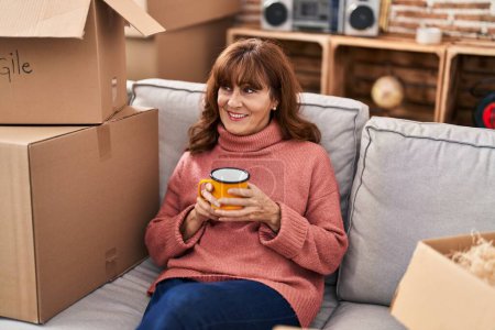 Foto de Middle age woman drinking coffee sitting on sofa at new home - Imagen libre de derechos