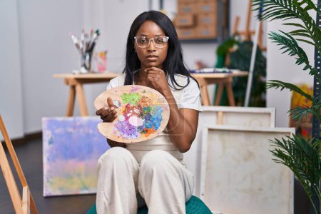 Téléchargez les photos : African young woman holding painter palette serious face thinking about question with hand on chin, thoughtful about confusing idea - en image libre de droit