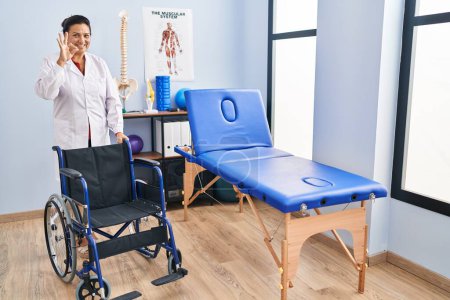 Foto de Mujer hispana de mediana edad con silla de ruedas en la clínica de fisioterapia haciendo signo bien con los dedos, sonriendo gesto amistoso excelente símbolo - Imagen libre de derechos