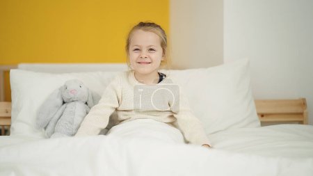 Téléchargez les photos : Adorable fille blonde souriante confiante assise sur le lit dans la chambre - en image libre de droit
