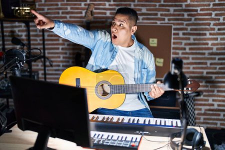 Photo for Hispanic young man playing classic guitar at music studio pointing with finger surprised ahead, open mouth amazed expression, something on the front - Royalty Free Image