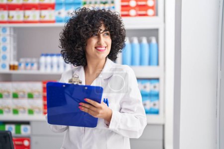 Téléchargez les photos : Young middle east woman pharmacist smiling confident writing on document at pharmacy - en image libre de droit