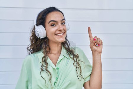 Téléchargez les photos : Jeune femme hispanique écoutant de la musique à l'aide d'écouteurs souriant avec une idée ou une question pointant du doigt avec un visage heureux, numéro un - en image libre de droit